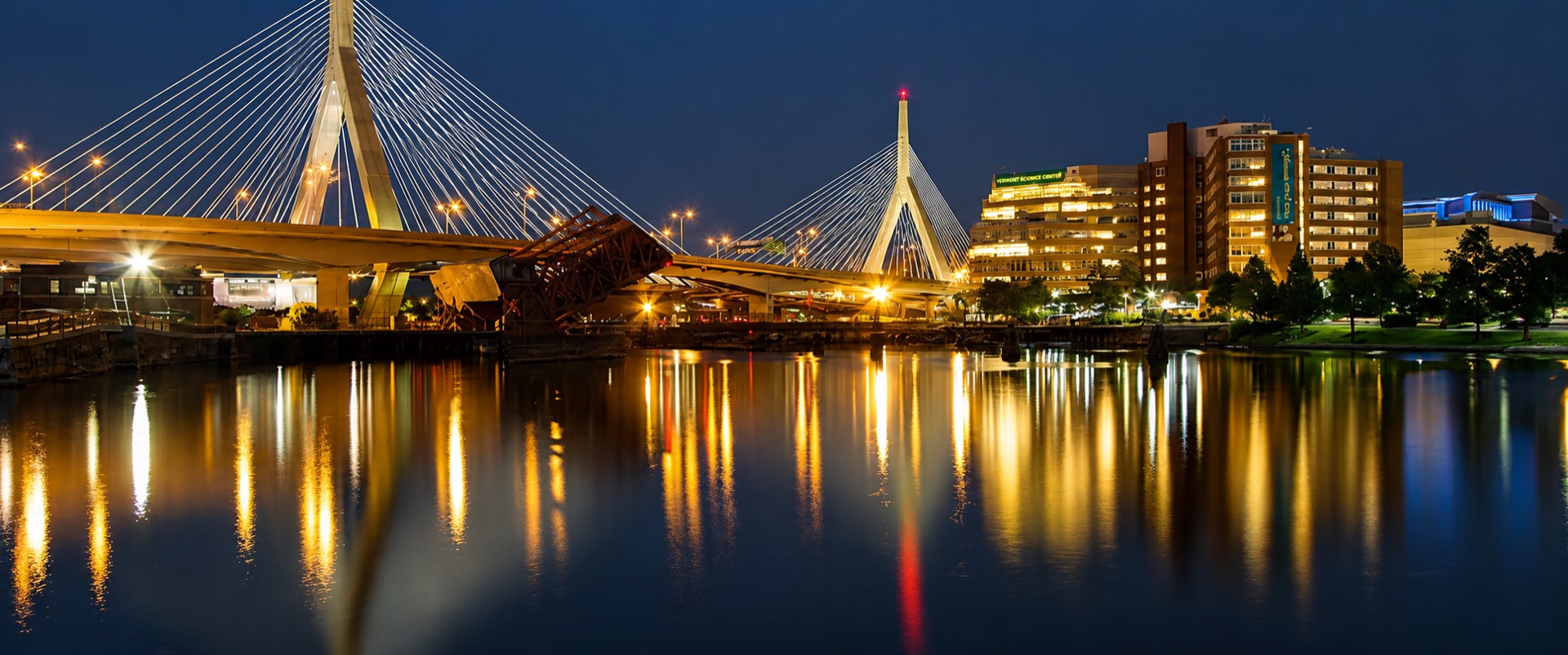 A New England city skyline at dusk with a cable-stayed bridge reflected in the river below.