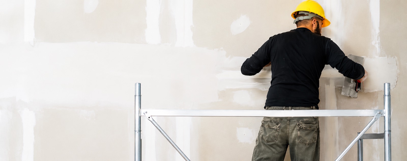 A drywall finisher on a rolling scaffold skim-coating a long commercial wall with a wide taping knife.