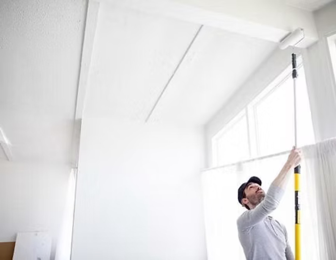 A painter using an extension pole to roll fresh white paint across a vaulted interior ceiling.