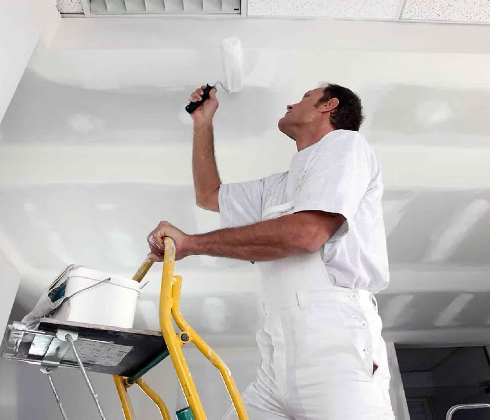 A painter in white overalls rolling a fresh coat of paint across a smoothly finished ceiling.