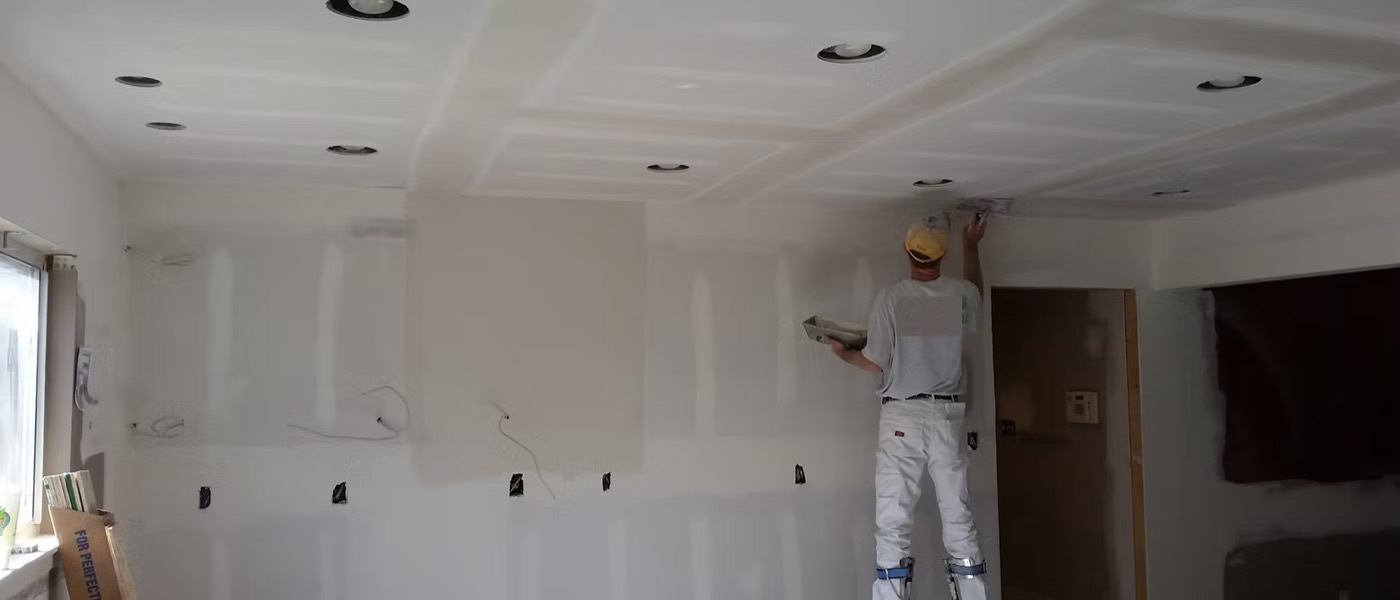 A finisher on stilts skim-coating a ceiling around recessed light openings.