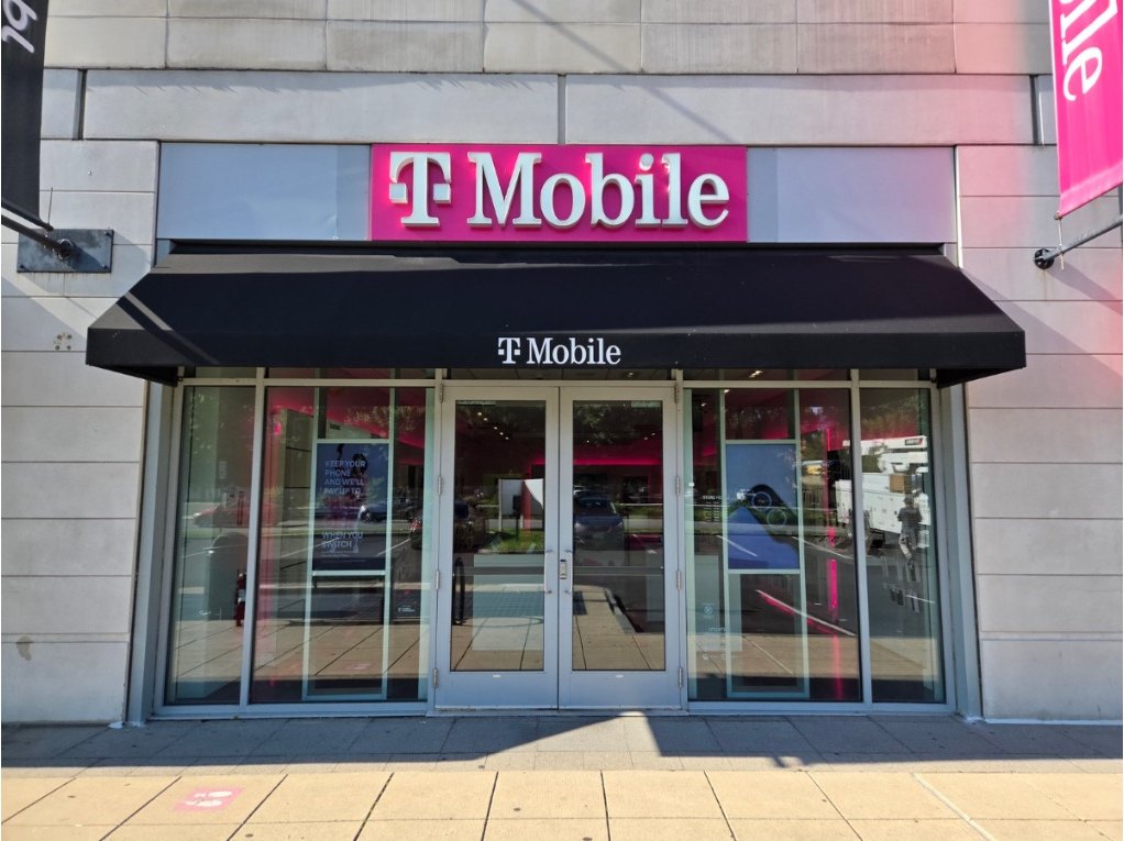 The T-Mobile storefront on Revere Beach Parkway in Medford, Massachusetts — an illuminated magenta sign set into a finished bulkhead above a black awning, with full-height glass doors and finished stone piers either side.