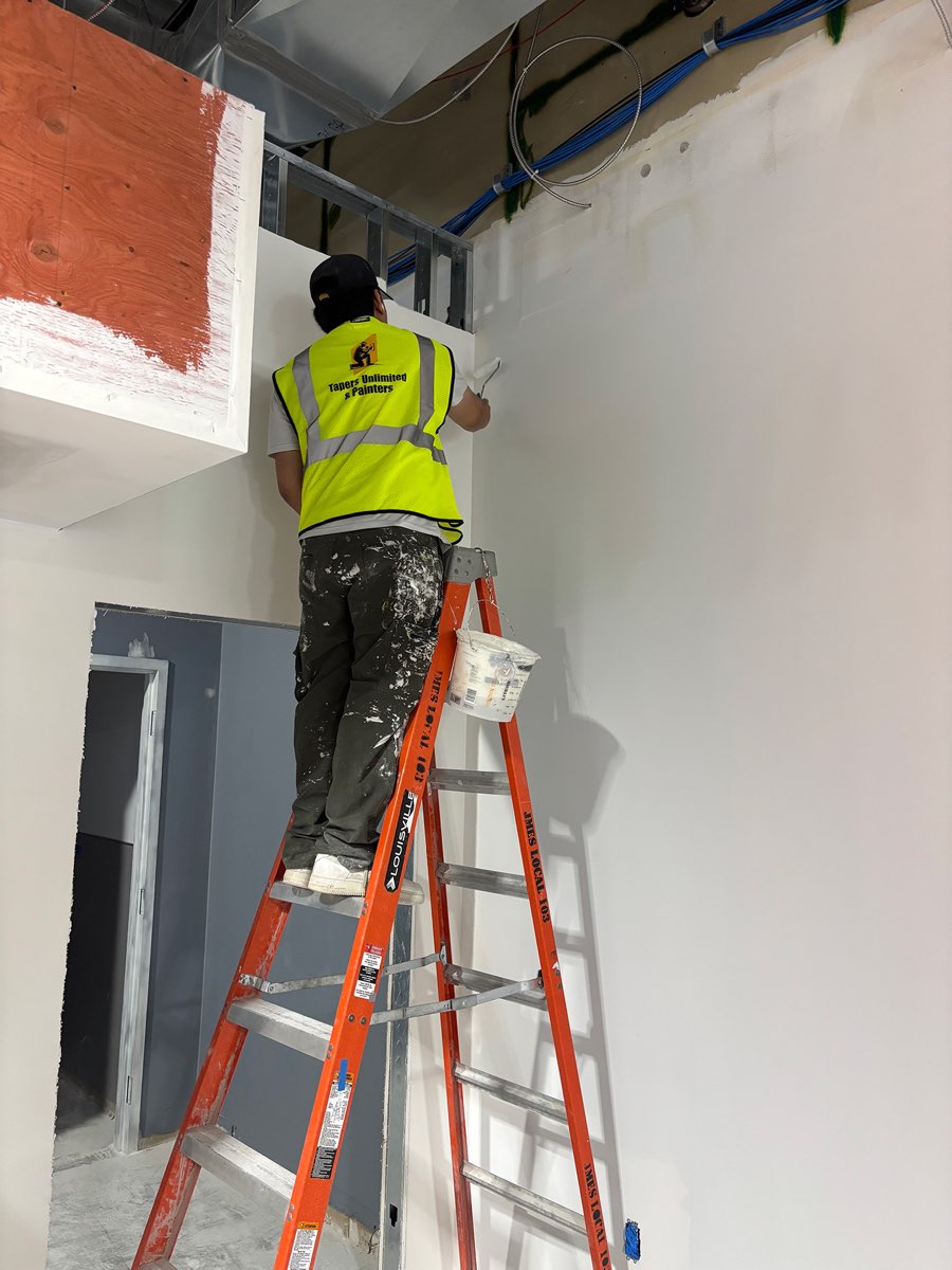 A Tapers Unlimited & Painters crew member in a branded hi-vis vest working from a step ladder to cut in paint at the head of a tall commercial wall, with a hanging paint kettle and the open services ceiling above.