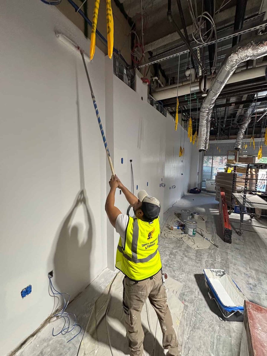 A Tapers Unlimited & Painters crew member in a branded hi-vis vest running a roller on a long extension pole up a tall, smoothly finished commercial wall, with the open services ceiling and caution tape above him.