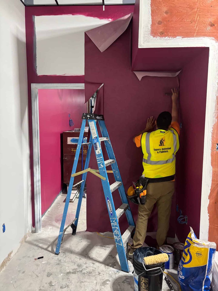 A Tapers Unlimited & Painters crew member in a branded hi-vis vest smoothing a length of wallpaper onto a deep magenta wall in a commercial fit-out, with a stepladder, paint buckets and joint compound on the floor beside him.